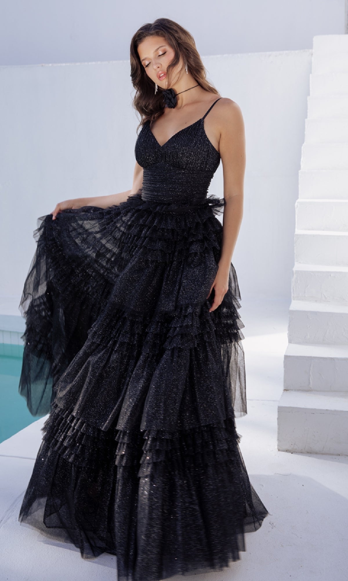 A woman in the Lace-Up Long Glitter-Tulle Prom Dress: Eureka 9888 stands by a white staircase and pool, wearing the sleeveless, tiered black gown and holding part of her dress with one hand.